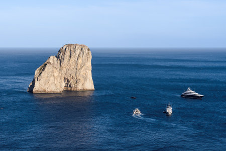 Boats navigate the azure waters near the iconic Faraglioni rocks off Capri Island during a clear day, showcasing Italy's stunning coastal beauty.の写真素材