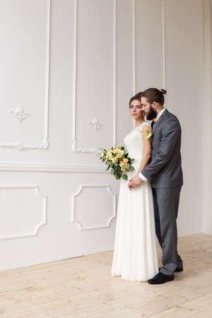 Beautiful bride in white dress with bouquet and handsome groom with beard in grey suite standing and embracing in bright interiorの写真素材