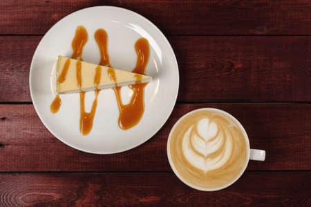 A cup of cappuccino and a piece of cheesecake, drizzled in caramel sauce are in white dish on wooden maroon table. The view from the topの写真素材