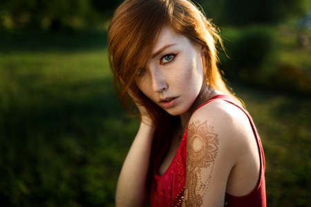 Close-up portrait of a redheaded young girl with a tattoo on her shoulder against a green background in the sunset lightの写真素材