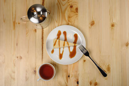 A plate of cheesecake with caramel sauce and a fork, a cup of tea and a teapot stand on a light wooden table. Top viewの写真素材