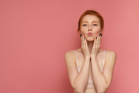 Redhead young woman wearing lace bra with a bun on head makes a kissy face on pink background