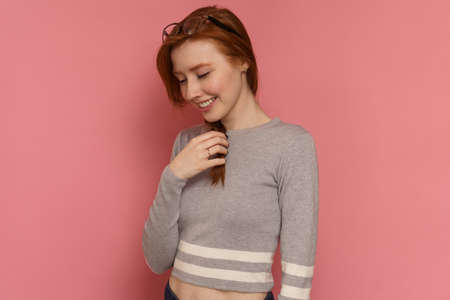 Red-haired young woman stands sideways to the camera, touches her braid and smiles on pink backgroundの写真素材