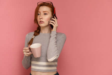 Redhead young woman with pink cup in one hand speaks on phone, pink backgroundの写真素材