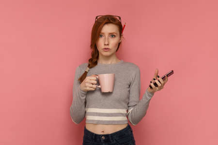 Portrait of a redhead girl with a cup in one hand and her smartphone in other handの写真素材