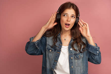 Young dark-haired curly woman listens to music with headphones looking up with her mouth open, pink backgroundの写真素材