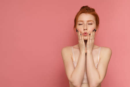 Young red-haired woman wearing lace bra makes a kissy face with her eyes closed, pink backgroundの写真素材