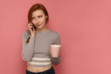 Red-haired girl with a cup listens to someone on her smartphone with funny face expressionの写真素材