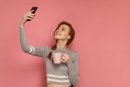 Redhead young woman smiles and poses with a cup in one hand while taking selfie