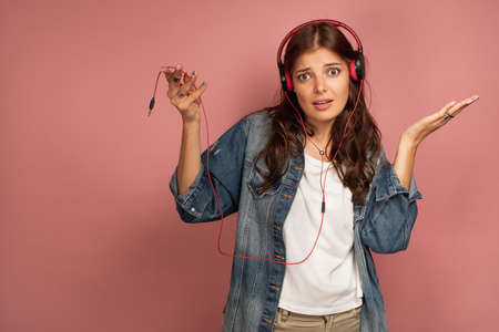 Young dark-haired confused woman spreads her hands out as her headphones are not connected to music device, pink backgroundの写真素材