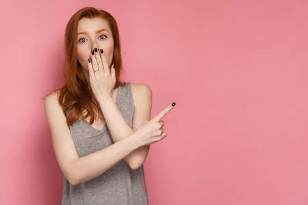 Redhead woman points her finger at blank space on the right of the photo covering her mouth with her hand in amazementの写真素材