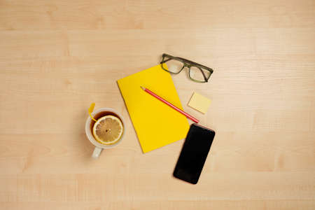 Yellow notepad with a red pencil, phone, glasses and a cup of tea are lying on a light wooden table.の写真素材
