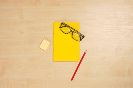 A yellow notebook with glasses, a red pencil and a sticker are in the middle of a light wooden...