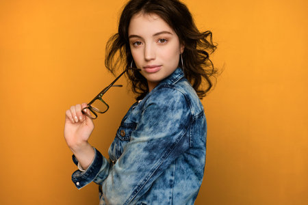 A curly dark-haired girl in a jeans stands on a yellow background in a half-turn and looks at the...