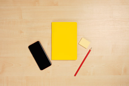 Yellow notepad, red pencil and phone lie in the middle of a light wooden table.