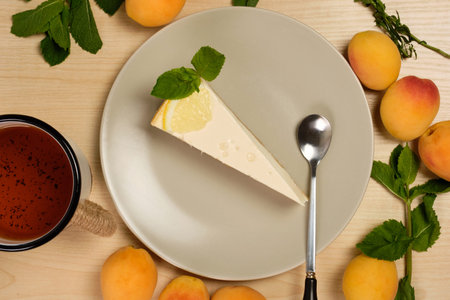 Cheesecake decorated with mint on a plate with a cup of tea stands surrounded by herbs and apricots...