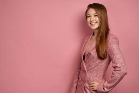 Red-haired girl in a pink jacket stands on a pink background and smiling looks at the camera standing half a turn.の写真素材
