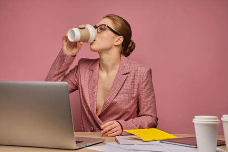 Redhead girl in a business suit and glasses on a pink background sitting at a table with papers and a laptop and drinking coffee.の写真素材