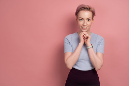 A girl with short pink hair in a T-shirt stands on a pink background, looking at the camera and...