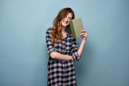 A young girl in a plaid shirt stands on a blue background hiding behind a book.の写真素材