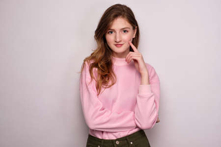 A girl in a pink sweater stands on a white background, looks at the camera, propping her cheek with her finger.の写真素材