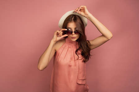 A dark-haired girl in a pink dress stands on a pink background, lifting her hat and skipping sunglasses, looking down to the side.の写真素材