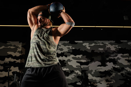 Muscular man in a sports uniform and a baseball cap stands holding the med ball above him and looks...