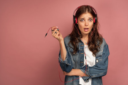 A dark-haired girl in jeans stands on a pink background in headphones and annoyedly holds the cord...