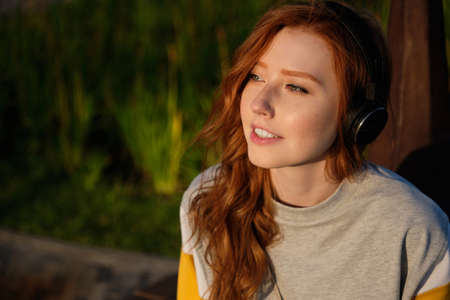 Portrait of a red-haired girl with freckles, green eyes in headphones, sitting in the sunset light and looking into the distanceの写真素材