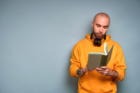 A bearded guy in a yellow sweatshirt stands on a blue background with headphones on his neck, looks...