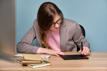 A girl in a pink sweater, gray jacket and glasses sits at a table, bowing her head, draws on an...