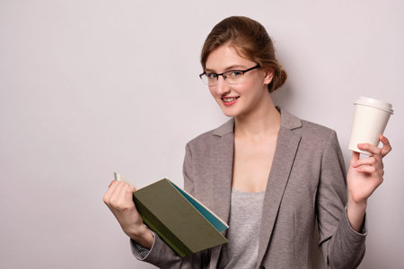 Blonde girl in glasses and a gray jacket with a book in his hands and a paper cup on a white...