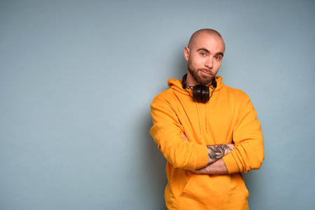 A bearded guy in a yellow sweatshirt stands with headphones on his neck, arms crossed over his chest and looks into the frameの写真素材