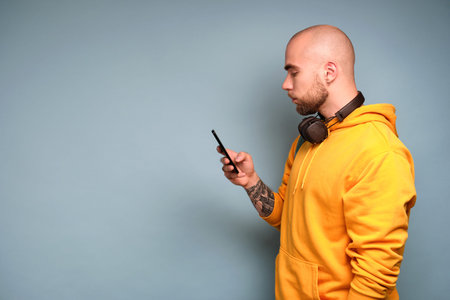 A bearded guy in a yellow sweatshirt stands in profile on a blue background with headphones on his...