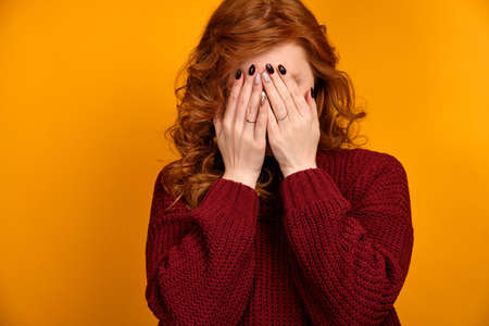 Red-haired curly girl in a burgundy sweater on a yellow background is standing covering her face with her handsの写真素材