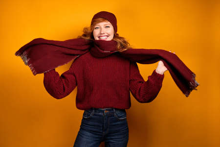 Red-haired curly girl in a burgundy sweater, scarf and hat looks at the camera smiling broadly against a yellow background.の写真素材