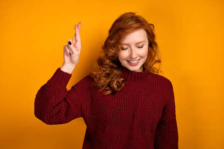 Beautiful red-haired girl in a burgundy sweater stands with her head bowed, smiling and raising her hand with crossed fingers.の写真素材
