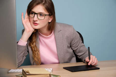 A girl in a gray jacket and glasses sits at a table and carefully looks at the monitor, working on a graphics tablet.の写真素材