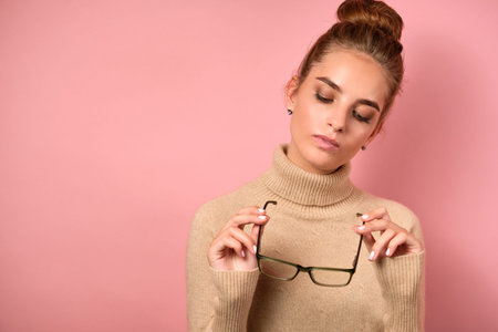 A girl in a beige turtleneck with gathered hair stands on a pink background and looks at the glasses...