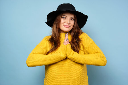 A beautiful brunette in a yellow sweater and black hat stands on a blue background, clasped her...