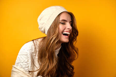 Portrait of a laughing girl in a white sweater and hat standing half-turn on a yellow background.の写真素材