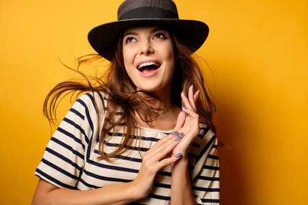 A beautiful brunette in a striped T-shirt and wicker hat is smiling on a yellow background, looking to the top with flying hairの写真素材