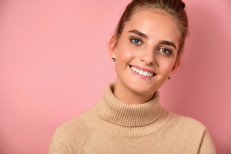 A girl with a high beam stands in a sweater against a pink background and smiles into the frame.