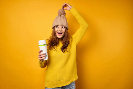 A beautiful brunette in a yellow sweater pulls the hat up by the pompom and looks to the side, holding a thermomugの写真素材