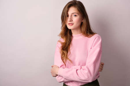 A fair-haired girl in a pink sweatshirt stands on a white background and hugs herself with her hands, looks into the frameの写真素材