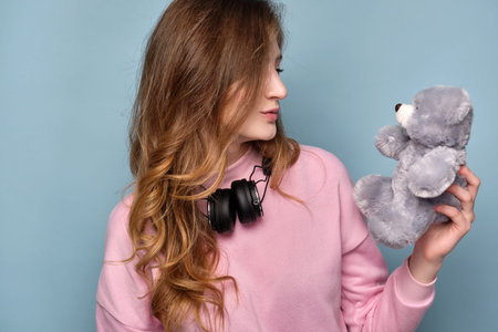 A girl in a pink sweatshirt with headphones on neck stands on a blue background in a half-turn and...