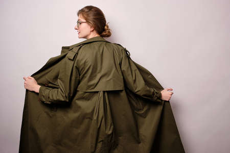 A girl stands on light background with her back to the frame, turning her head in profile and opening the green trench to the sideの写真素材