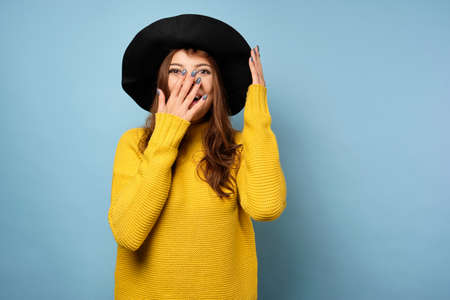 Brunette in a yellow sweater stands on a blue background, straightening his black hat and laughing, covering his face with handの写真素材