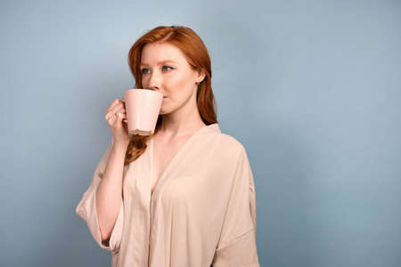 A red-haired girl in a light robe stands half a turn on a blue background and takes a sip from a light mug.の写真素材