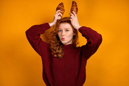 A red-haired curly girl in a burgundy sweater stands on an orange background and puts croissants to her head, stretching her lipsの写真素材